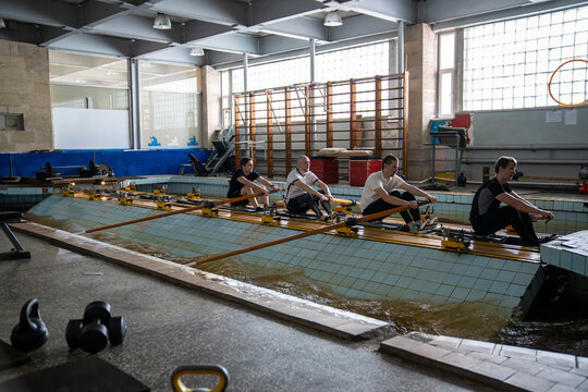 Four male athletes pull long wooden oars from fixed machines beside indoor pool. Synchronized effort combines strength, endurance and concentration during physically demanding team workout.