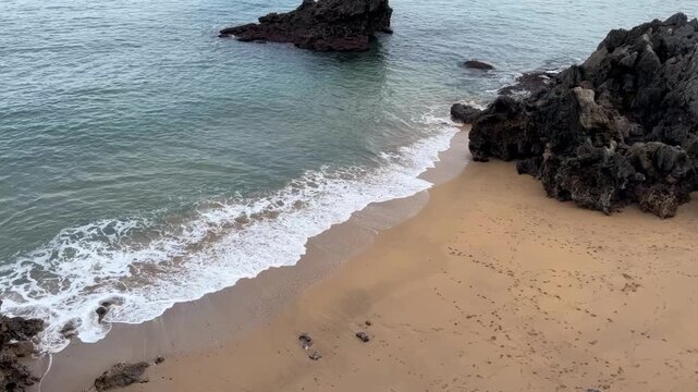 High-angle video footage of gentle ocean waves rolling onto a golden sandy beach flanked by dark volcanic rocks. The turquoise sea water meets the shore, leaving footprints and white foam patterns in 