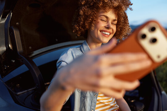 Selfie of a young woman with curly hair smiling in a denim jacket and striped shirt inside a car, sunlight on her face for a bright casual moment.