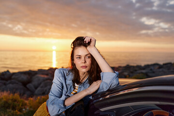 A young woman leans on a car at sunset by the rocky coastline, wearing a denim jacket. A calm, reflective moment ideal for lifestyle, travel, and self care themes. © SHOTPRIME STUDIO