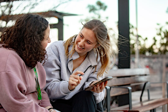 Two young women engaged in conversation while sitting on an urban bench looking at a smartphone