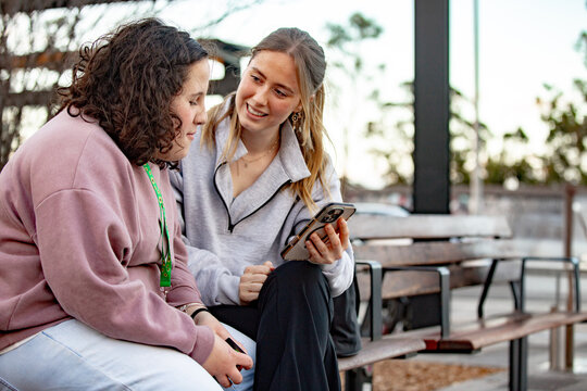 Two young women engaged in conversation while sitting on an urban bench looking at a smartphone