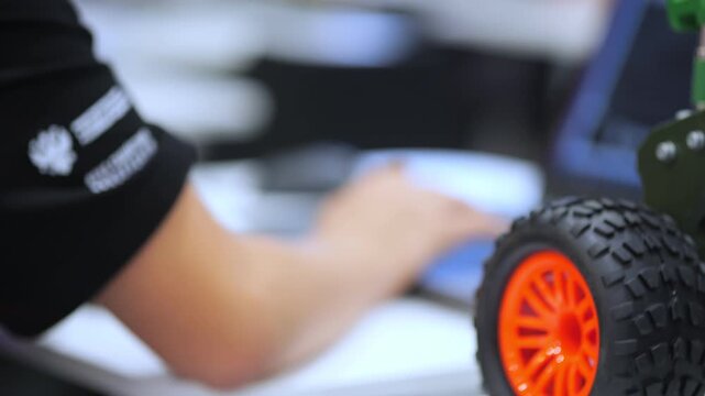 Person works with robot on desk. Orange wheel details stand out. Focus on mechanical parts. Background blurred for detail. Tech workspace scene
