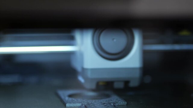 3D printer nozzle moves slowly over build platform. Focused shot of extruder head depositing material layer by layer. Dark background with soft blue light highlights mechanical motion