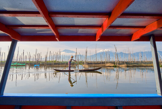 View of a fisherman in a traditional wooden boat on Lake Rawa Pening, with bamboo fishing poles and calm water, mountains in the distance, Waduk Rawapening, Central Java, Indonesia.