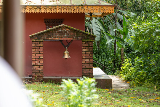 View of the Bangladi Shiva Temple featuring a traditional brass bell hanging from a brick structure, surrounded by lush green foliage in Pokhara, Gandaki Province, Nepal.