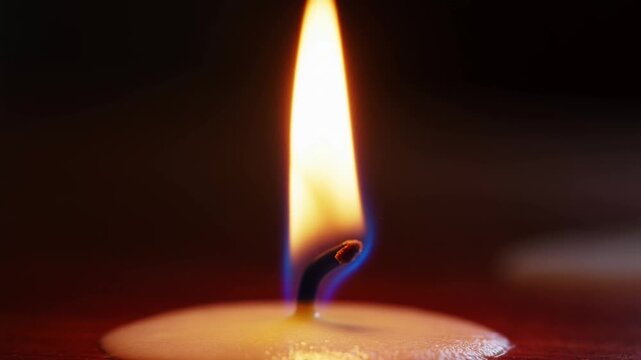 Slow motion static close up of candle flame flickering and wick bending as wax melts on tabletop in dim room