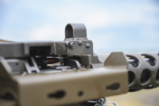 Detailed close up of a mounted heavy machine gun showing the metal iron sight, receiver and perforated barrel assembly against a softly blurred outdoor background and blue sky.