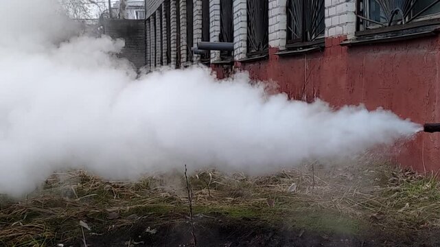 Heavy white smoke and steam escaping from a basement drainage pipe of an old brick public building with windows, depicting a city infrastructure utility failure and emergency accident.