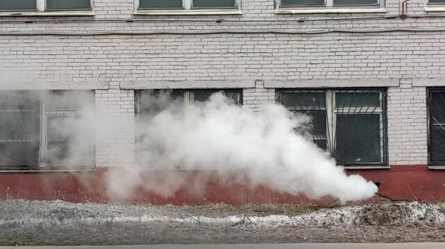 Heavy white smoke and steam escaping from a basement drainage pipe of an old brick public building with windows, depicting a city infrastructure utility failure and emergency accident.