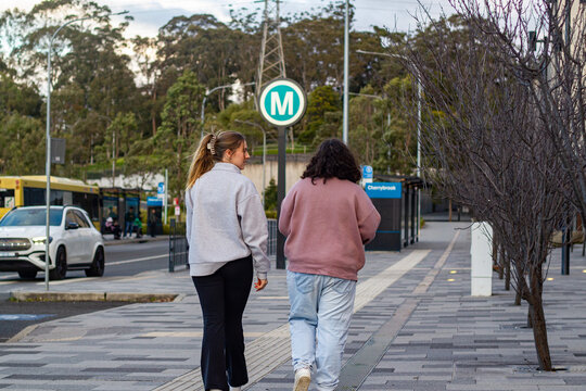 Two young women walking side by side along a paved footpath near a metro station