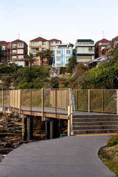 Coastal pedestrian walkway and stairs leading past rocky vegetation toward modern apartments