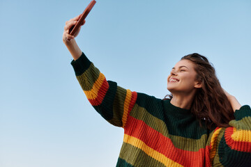 A smiling woman in a colorful striped sweater takes a selfie outdoors against a clear blue sky, capturing a playful moment and youthful confidence for lifestyle travel themes today. © SHOTPRIME STUDIO
