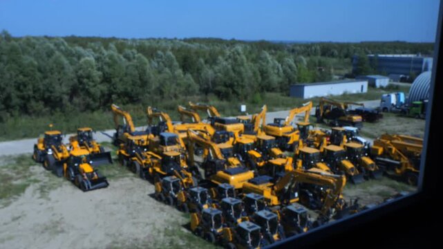 Large outdoor yard filled with many yellow construction machines. Machines stand in rows on dirt ground. Background shows green trees and clear blue sky. Buildings visible at far edge of scene
