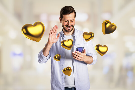 Smiling young man waving while holding a smartphone, surrounded by floating golden heart icons for dating and social media connection technology.