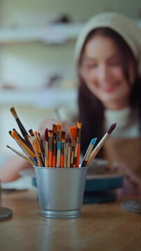 Close up of colorful paintbrushes in container with young Asian woman painting pottery in blurred background, creative ceramic workshop, art supplies, craftsmanship.