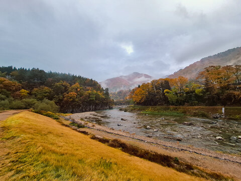 View of the river flowing through the autumn landscape with colorful trees and mountains under a cloudy sky in Shirakawa, Gifu, Japan.