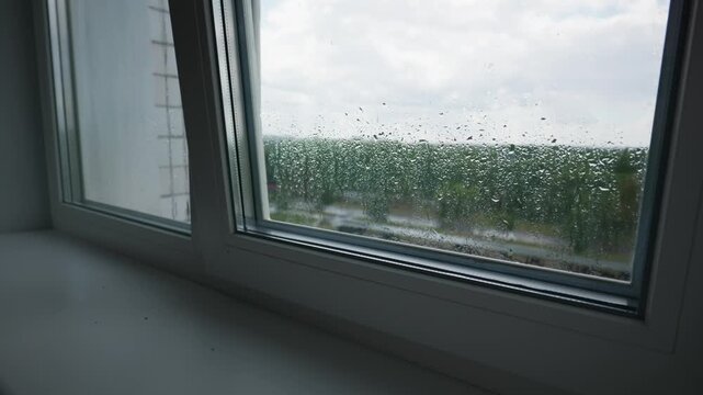 Water droplets accumulate on a double-glazed modern window pane overlooking green trees. The white vinyl frame and sill showcase durable construction against outdoor wet weather.