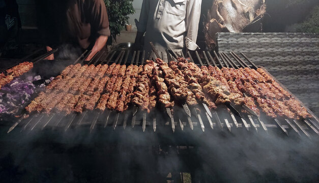 View of a large outdoor grill loaded with traditional seekh kebabs and tikka boti cooking over hot coals with smoke rising in Multan, Punjab, Pakistan.