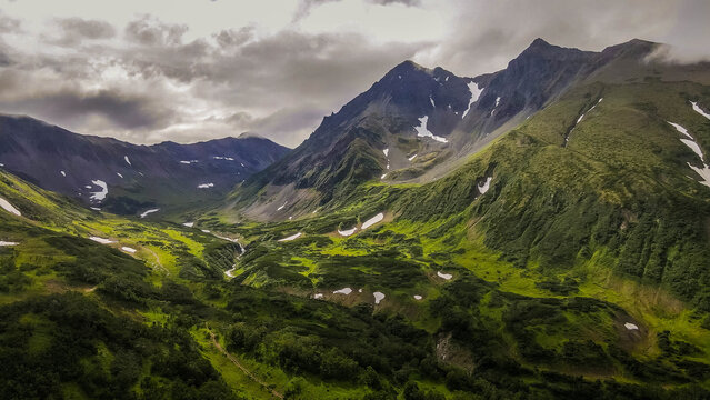 Beautiful aerial view of the Vachkazhets mountain massif in Kamchatka, revealing dramatic glacial cirques, lush green valleys, alpine meadows, rocky peaks, and a clear mountain lake.
