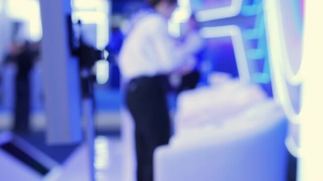 Blurred figures in modern, blue-lit indoor space. People stand near white furniture and glowing structures. Background features abstract, illuminated panels