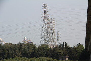 Obraz premium High voltage power transmission towers and pylon electricity lines near industrial power plant complex behind green trees