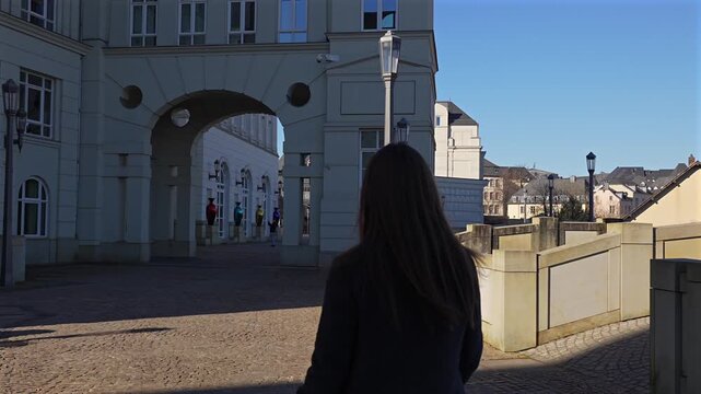 Rear view of a woman walking along a sunlit elevated terrace overlooking the rooftops and old town of Luxembourg