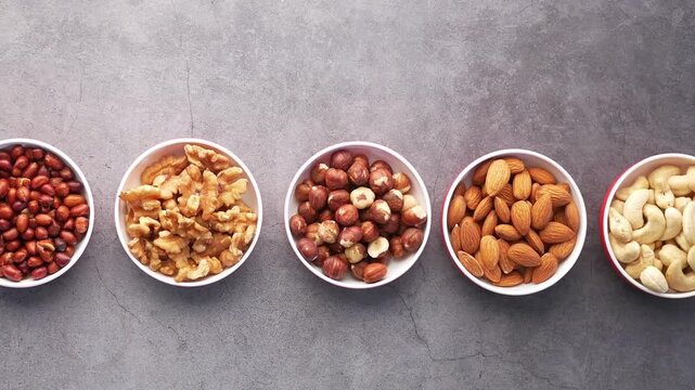 Top view of various nuts in white bowls sliding across grey background in slow motion
