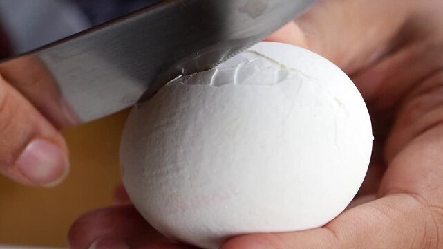 Close up video of hands holding a white egg with cracks and a knife slowly peeling off the shell