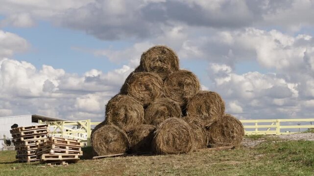 round hay bales are stacked in a pyramid beneath dramatic summer clouds, presenting harvested livestock feed, rural agriculture, seasonal work and food security
