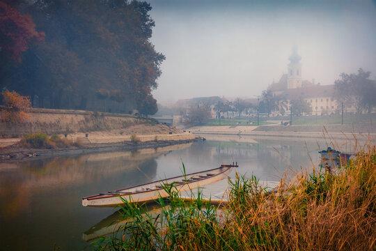 Foggy morning view of popular tourist attraction - Regi csonakhaz with Church of Our Lady of Mount Carmel on background. Nice autumn cityscape of Gyor town, Hungary, Europe. 