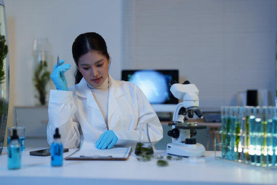 Female scientist working late in a research laboratory, wearing gloves and a lab coat, analyzing samples and taking notes with a microscope and test tubes on the table
