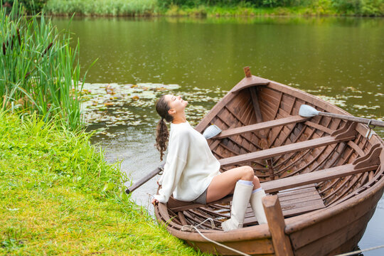 Young woman relaxing in wooden rowboat by lake on autumn day. Cozy sweater and rubber boots represent seasonal outdoor lifestyle and slow living in nature