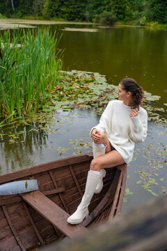Young woman relaxing in wooden rowboat by lake on autumn day. Cozy sweater and rubber boots represent seasonal outdoor lifestyle and slow living in nature