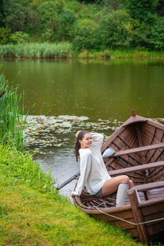 Young woman relaxing in wooden rowboat by lake on autumn day. Cozy sweater and rubber boots represent seasonal outdoor lifestyle and slow living in nature