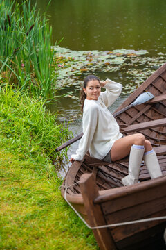 Young woman relaxing in wooden rowboat by lake on autumn day. Cozy sweater and rubber boots represent seasonal outdoor lifestyle and slow living in nature