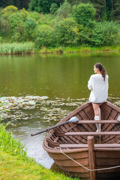 Young woman in white sweater stands on wooden boat by calm lake surrounded by lush greenery and lily pads, enjoying serene nature view in summer