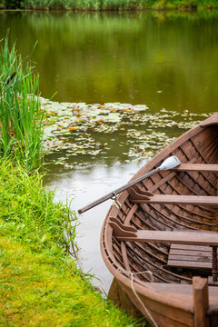 Wooden rowboat with oars resting on calm water surrounded by green grass and lily pads near a wooden dock in a serene natural setting