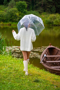 Young woman in white sweater and knee-high socks walks with a clear umbrella by a tranquil lake, wooden boat moored nearby, surrounded by lush greenery