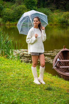 Young woman in white sweater and rubber boots holding a clear umbrella stands on green grass by a tranquil lake with a wooden boat in the background