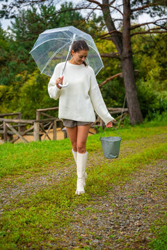 Young woman in white sweater and rubber boots holding a bucket and clear umbrella walking on a gravel path near a lake surrounded by greenery