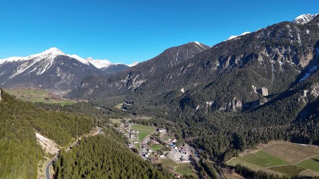 albula valley alpine panorama switchback mountain road farmland fields village rooftops forested slopes snowcapped peaks graubunden switzerland aerial drone footage wide shot clear sky springtime