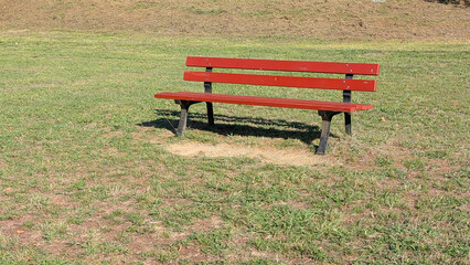 A red wooden park bench with black metal legs stands alone on a grassy field. © Beautiful talkers