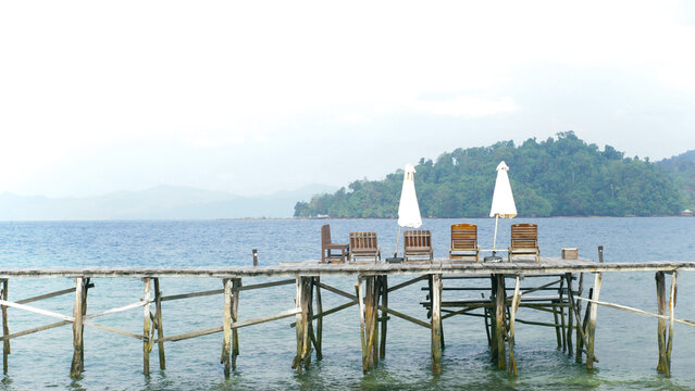 View of a wooden pier with lounge chairs and umbrellas overlooking the sea and lush tropical islands in Raja Ampat, Southwest Papua, Indonesia.