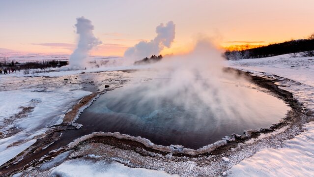 View of Strokkur Geyser erupting with steam rising into the golden sky above a snow-covered geothermal landscape in Selfoss, Blaskogabyggd, Iceland.