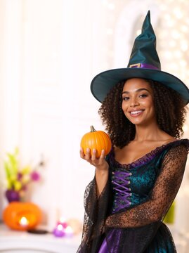 Smiling young Black woman in witch costume holding a small pumpkin. Happy African American female dressed up for Halloween party. Bright indoor background with festive lights