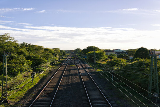 Elevated view of electrified railway tracks and switches running through a green suburban neighborhood beneath a bright blue sky.