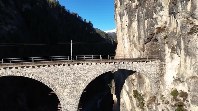 stone railway viaduct curves along sheer limestone cliff near filisur graubunden switzerland arches span forested ravine distant snowcapped peak aerial drone footage clear blue sky alpine gorge
