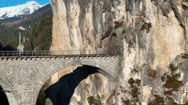 tunnel portal carved into sheer limestone cliff marks landwasser viaduct end near filisur switzerland arches curve along cliff base snowcapped peak aerial drone footage clear sky alpine gorge