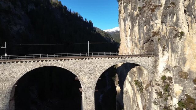 albula line viaduct arches curve toward rock cliff near filisur switzerland vegetation clings to limestone ledges forested gorge snowcapped mountain aerial drone footage clear sky alpine scenery